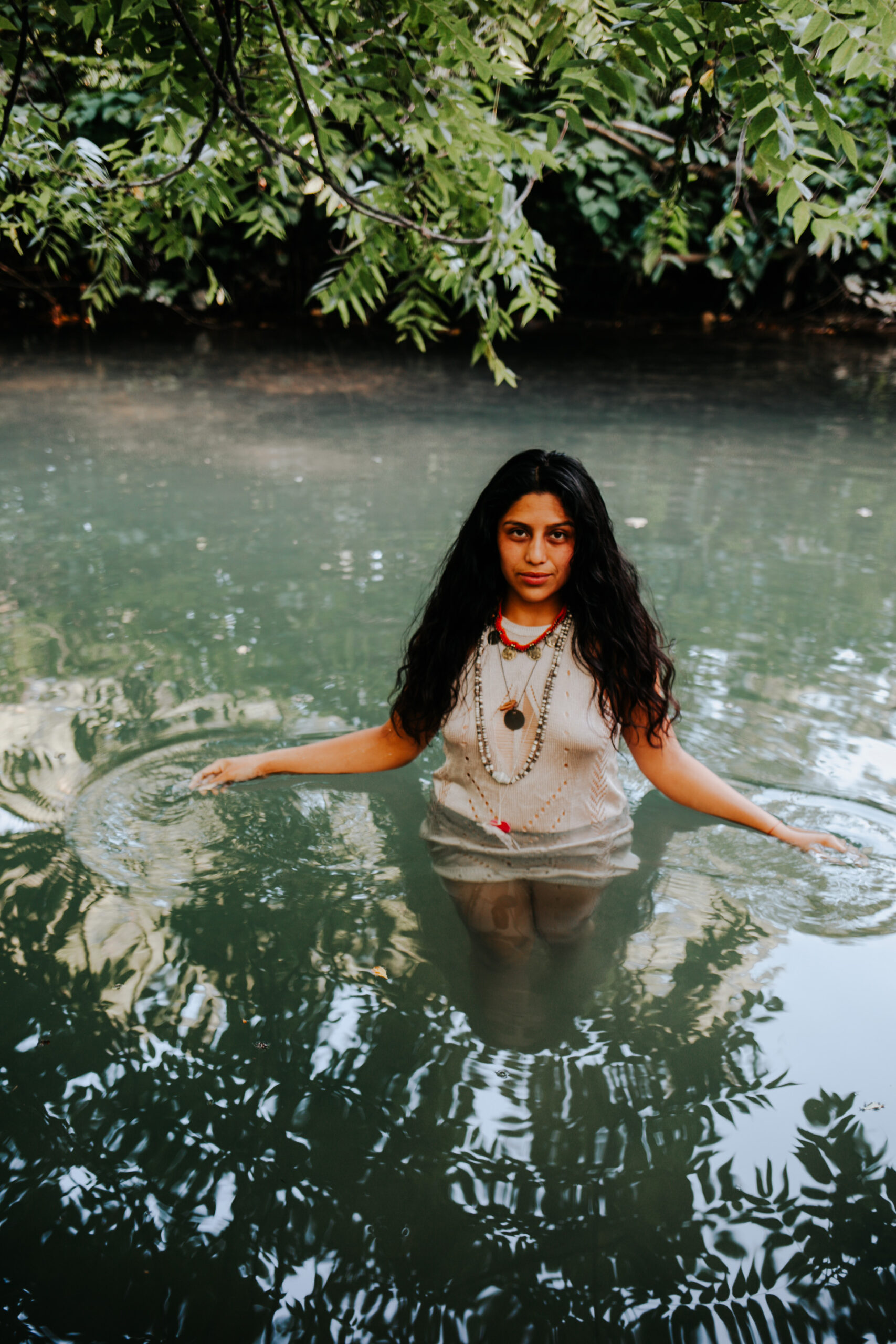 Leslie Avila, wearing white dress and necklaces, standing in waste-high water, greenery in background and reflected in the water, her out hands to her sides gently moving the water